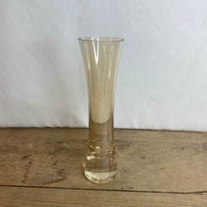 Può includere: Un vaso alto e sottile in vetro color ambra. Il vaso ha una parte superiore svasata e una base stretta. È fatto di vetro trasparente e poggia su una superficie di legno.