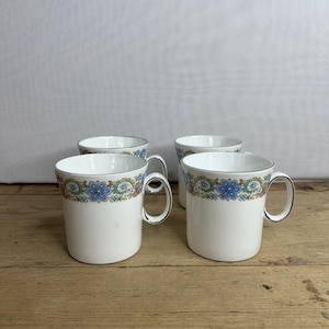 May include: Four white ceramic mugs with a floral design in blue, green, and gold. The mugs have a silver trim on the rim and handle. The mugs are on a wooden surface.
