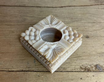 May include: A square, cream-colored, carved stone candle holder with a circular opening in the center. The holder has a detailed design of grapes and leaves around the opening.