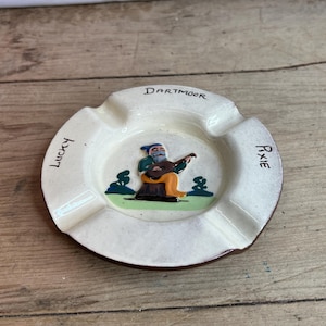 Puede incluir: Un cenicero de cerámica blanca con un borde marrón. Presenta una ilustración de dibujos animados de un duende tocando la guitarra, con las palabras "Dartmoor Pixie" escritas alrededor del borde.