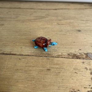 May include: A small glass turtle figurine. It has a brown shell and blue flippers and head. The turtle is on a wooden surface. The figurine is a decorative item.