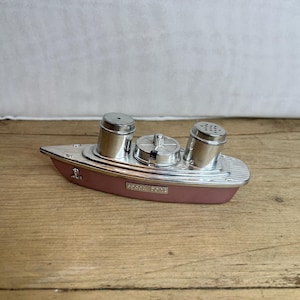 May include: A vintage boat-shaped salt and pepper shaker set. The base is a muted rose color with silver-toned accents. Two cylindrical shakers and a central compartment are visible. The words "Speed Boat" are printed on the side.