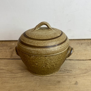 Może przedstawiać: Beżowa ceramiczna miska z pokrywką, o zaokrąglonym kształcie i dwóch małych uchwytach. Pokrywka posiada pętlę i jest ozdobiona ciemnobrązowymi poziomymi paskami. Miska ma nakrapianą fakturę i stoi na drewnianej powierzchni.