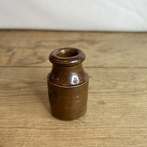 Peut inclure: Petite bouteille en grès brun avec un col étroit et un bord large et arrondi. Le corps de la bouteille est cylindrique avec une bande blanche au milieu. La bouteille est posée sur une surface en bois.