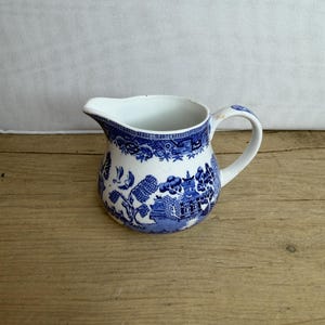 Könnte beinhalten: Eine weiße und blaue Keramik-Sahnekännchen mit Henkel und Ausguss. Das Sahnekännchen hat ein blaues Willow-Muster mit Bäumen, Gebäuden und einer Brücke. Das Sahnekännchen steht auf einer Holzoberfläche.