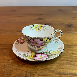 Puede incluir: Un juego de taza y platillo vintage. La taza y el platillo de porcelana blanca están decorados con diseños florales rosas, amarillos y marrones. La taza tiene un borde y un asa dorados. El platillo también tiene un borde dorado. El juego está sobre una superficie de madera.