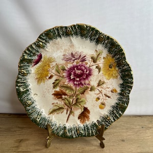 Pode incluir: Um prato decorativo com um design floral. O prato apresenta um centro creme com um buquê de flores roxas, amarelas e brancas. A borda é verde com detalhes dourados. O prato é exibido em um suporte dourado.