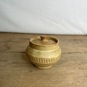 May include: A small, round, beige ceramic jar with a lid. The jar has a textured, geometric pattern around the middle and a small knob on top of the lid. The jar is sitting on a wooden surface.