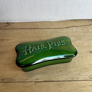 May include: A green ceramic hair pin box with the words "HAIR PINS" in a stylized font. The box has a rectangular shape with rounded edges and a glossy finish. The box is sitting on a wooden surface.