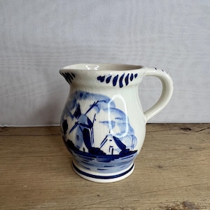 Puede incluir: Jarra de cerámica blanca con un diseño de molino de viento azul. La jarra tiene un asa y un borde acampanado con detalles azules. La base también está acentuada con azul. La jarra está sobre una superficie de madera.