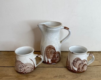 May include: A white ceramic pitcher and two mugs with brown abstract designs. The pitcher has a handle and spout. The mugs and pitcher are on a wooden surface. The mugs have handles.