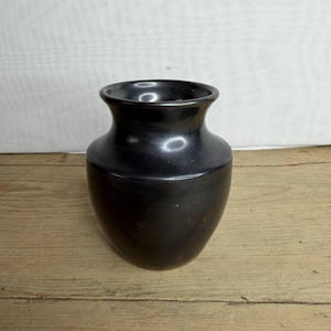 Könnte beinhalten: Eine schwarze Keramikvase mit rundem Körper und leicht ausgestelltem Rand. Die Vase hat eine glatte, glänzende Oberfläche und steht auf einer Holzoberfläche. Die Vase ist leer und kann mit Blumen gefüllt oder als Dekoration verwendet werden.