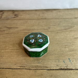May include: A small, hexagonal trinket box with a green and white floral design. The lid features a detailed pattern of gold, blue, and white flowers on a green background. The box is set on a wooden surface.