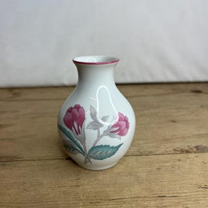May include: A small, white ceramic vase with a floral design. The vase has a red rim and is decorated with pink flowers, grey leaves, and teal accents. The vase is approximately 10 cm tall.