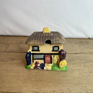 May include: A miniature ceramic cottage with a thatched roof, yellow chimney, and a red door. The cottage has black window frames and is surrounded by colorful landscaping, including blue, red, and yellow accents. The cottage sits on a wooden surface.