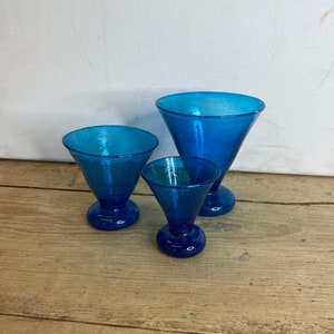 Könnte beinhalten: Drei blaue Martini-Gläser in verschiedenen Größen. Jedes Glas hat eine strukturierte Oberfläche, eine konische Schale und einen abgerundeten Fuß. Die Gläser sind auf einer Holzoberfläche angeordnet.