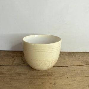May include: A cream-coloured ceramic bowl with a rounded shape. The exterior has a textured, striated pattern with subtle brown accents. The interior is a smooth, bright white. The bowl sits on a wooden surface.