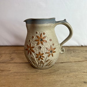 May include: A beige ceramic pitcher with a floral design. The pitcher has a curved handle and a spout. The flowers are brown with dark centers. The rim of the pitcher is blue. The pitcher is sitting on a wooden surface.