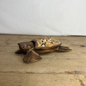 Pode incluir: Uma estatueta de tartaruga de madeira com uma carapaça de mosaico. A tartaruga é esculpida em madeira castanha escura, com uma carapaça feita de pequenas telhas de mosaico de cor clara. A cabeça e as barbatanas da tartaruga estão estendidas.