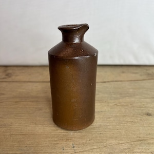 May include: A brown stoneware bottle with a narrow neck and a flared rim. The bottle has a cylindrical body and a slightly textured surface. The color is a warm, earthy brown, suggesting an antique or vintage item. The bottle is likely for decorative use.