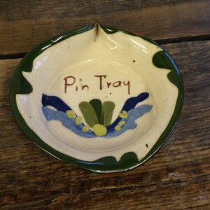 May include: Hand-painted ceramic pin tray with a scalloped green edge. The off-white interior features a floral design in blue, green, and yellow, with the words "Pin Tray" in brown script. A small hole is at the top for hanging.
