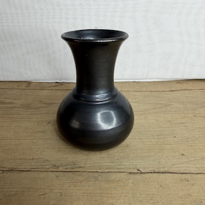 Op de afbeelding: Een kleine, zwarte vaas met een afgeronde body en een uitlopende hals. De vaas heeft een gladde, glanzende afwerking en staat op een houten oppervlak. De vaas is ongeveer 15 cm hoog.