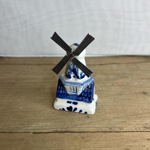 Puede incluir: Una figura de molino de viento de cerámica en miniatura. El molino de viento presenta un cuerpo blanco con patrones florales y geométricos azules. Las aspas son de color gris oscuro. La base es blanca con detalles florales azules. Sobre una superficie de madera.