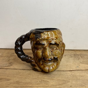 Peut inclure: Une tasse en céramique marron en forme de visage souriant. La tasse a une anse marron foncé et un intérieur marron foncé. Le visage présente des traits détaillés, dont un nez, des yeux et une bouche.
