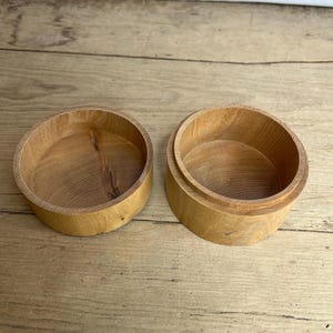 Puede incluir: Dos pequeñas cajas redondas de madera con tapas, expuestas abiertas. Las cajas son de madera de color claro, con patrones de vetas visibles. Las cajas están sobre una superficie de madera.