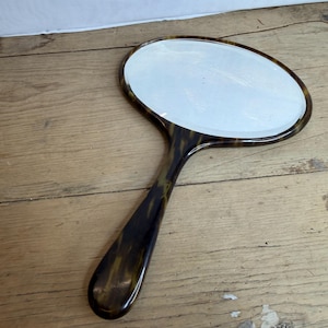 Op de afbeelding: Ovale handspiegel met een schildpadpatroon op het handvat en frame. De spiegel heeft een reflecterend oppervlak, tegen een houten achtergrond.