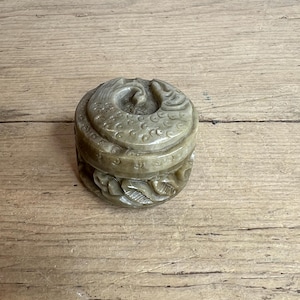 Puede incluir: Una pequeña caja redonda de piedra tallada con tapa. La caja es de color verde claro y presenta tallas detalladas de un dragón o serpiente en la tapa y patrones florales alrededor de los lados. Sobre una superficie de madera.