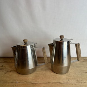 Puede incluir: Dos cafeteras de acero inoxidable con asas de madera. Las cafeteras tienen un acabado pulido y un diseño clásico.