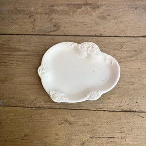 May include: A small, white, decorative dish with a scalloped edge and embossed floral details. The dish is oval-shaped and sits on a wooden surface. The dish is likely made of ceramic or porcelain.