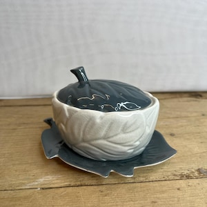 Op de afbeelding: Een keramische suikerpot met deksel en een schotel in de vorm van een blad. De kom en het deksel zijn donkerblauwgroen, terwijl de kom zelf gebroken wit is. Het ontwerp heeft een bladerenpatroon.