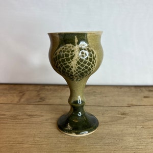 May include: A handmade ceramic goblet with a green and tan color scheme. The goblet features a detailed, textured heart design on the bowl. The base and stem are dark green, while the bowl has a lighter tan hue.