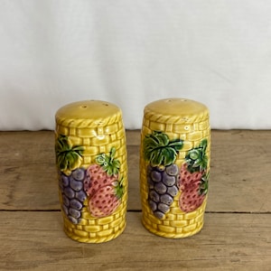 Puede incluir: Un par de saleros y pimenteros de cerámica. Los saleros y pimenteros son amarillos con un diseño de tejido de cesta. Cada salero y pimentero presenta un racimo de uvas moradas y una fresa. Las tapas tienen tres agujeros pequeños.