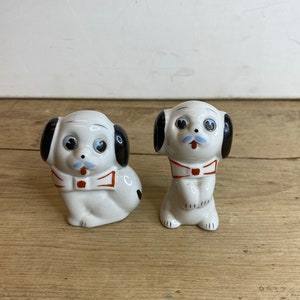 May include: Two ceramic dog figurines. The dogs are white with black ears, blue mustaches, and red bow ties. One dog is sitting, and the other is standing. The figurines are on a wooden surface.