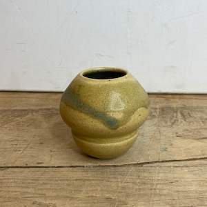 May include: A small, handmade ceramic vase with a rounded body and a narrow opening. The vase is a muted yellow-green color with a darker green band around the middle. The base is slightly wider than the top.
