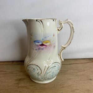 Puede incluir: Una jarra vintage de color crema con un patrón floral azul y blanco y detalles dorados. La jarra tiene un asa y una base redondeada.