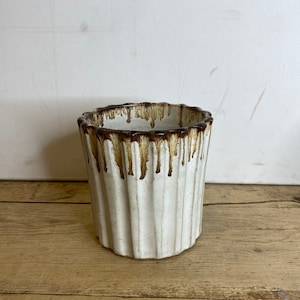 May include: A white ceramic planter with a ribbed design. The rim and upper portion of the planter have a brown, dripped glaze. The planter is cylindrical and sits on a wooden surface.