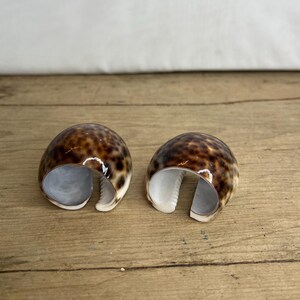 May include: Two natural seashell napkin rings. The shells have a brown and tan spotted pattern with a white interior. The napkin rings are open on one side, revealing the interior. They are displayed on a wooden surface.