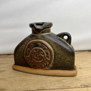 Può includere: Brocca in ceramica marrone con beccuccio e manico. La brocca presenta un emblema circolare con la parola "PAIN" al centro. La base è marrone chiaro e la brocca ha una superficie strutturata.