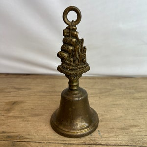 May include: A vintage brass bell with a detailed ship design on top. The bell has a loop for hanging and a flared base. The ship design includes intricate details of the ship's sails and structure. The bell is likely made of brass and has a tarnished appearance.