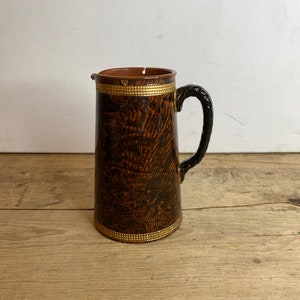 Puede incluir: Jarra antigua con exterior marrón oscuro texturizado y asa negra. La jarra está adornada con ribetes dorados en la parte superior e inferior. El interior es de color marrón claro. La jarra está sobre una superficie de madera clara.