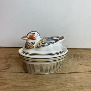 Puede incluir: Una figura de pato de cerámica con un diseño detallado. El pato tiene un cuerpo blanco, detalles en marrón y naranja, y está posado sobre un recipiente de estilo tejido. El recipiente tiene rayas negras y beige. El diseño general es decorativo.