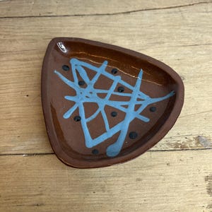 Puede incluir: Un plato de cerámica marrón con forma triangular. El plato está decorado con un diseño abstracto azul claro y pequeños puntos negros. El plato está sobre una superficie de madera.