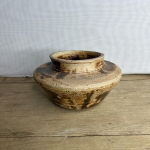 Op de afbeelding: Een handgemaakte keramische vaas met een brede, afgeronde vorm en een smalle, licht uitlopende rand. De vaas heeft een gespikkelde, aardse kleurenpalet van beige, bruin en bruin, met donkerbruine strepen. De vaas staat op een houten oppervlak.