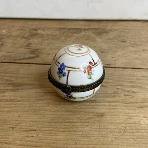 May include: A white, spherical trinket box with a hinged lid. The box is decorated with floral patterns in blue, pink, and red, accented with gold trim. The metal hinge and clasp are dark bronze.