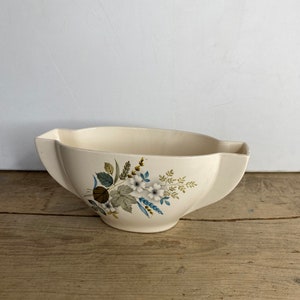 May include: Cream-colored, vintage ceramic serving bowl with a floral design. The bowl has a unique shape with flared sides and a decorative floral pattern in shades of blue, green, and white. The bowl is sitting on a wooden surface.