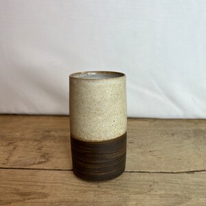 Op de afbeelding: Een handgemaakte keramische beker met een tweekleurig ontwerp. De bovenste helft is gespikkeld beige, terwijl de onderste helft donkerbruin is. De cilindrische beker staat tegen een neutrale achtergrond en toont zijn eenvoudige, moderne esthetiek.